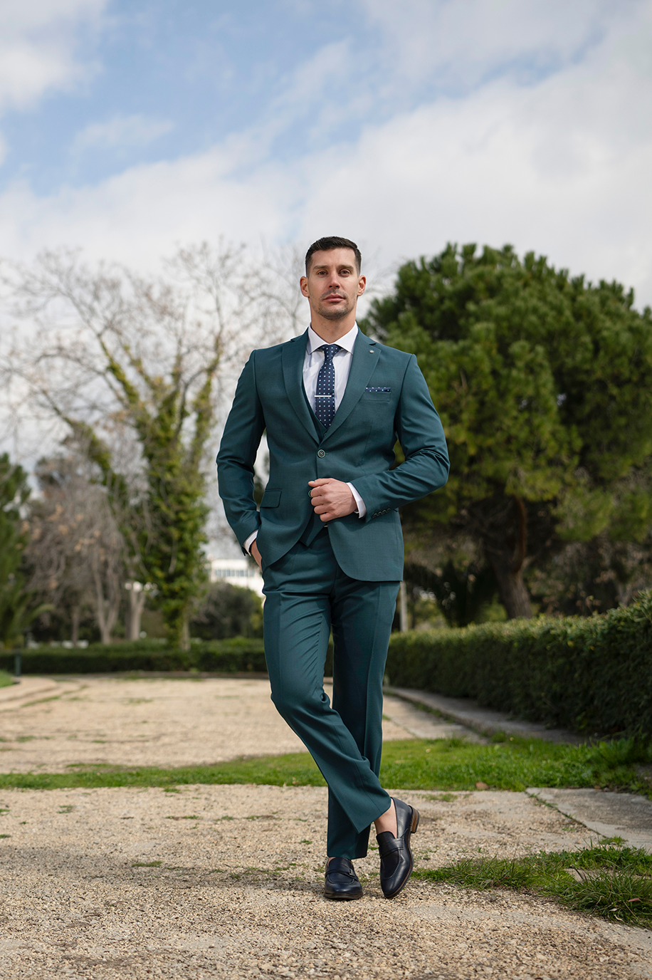 Ανδρικό κοστούμι Boston σε πράσινο. Formal εμφάνιση με loafers και σταυρωμένο πόδι σε φυσικό τοπίο.